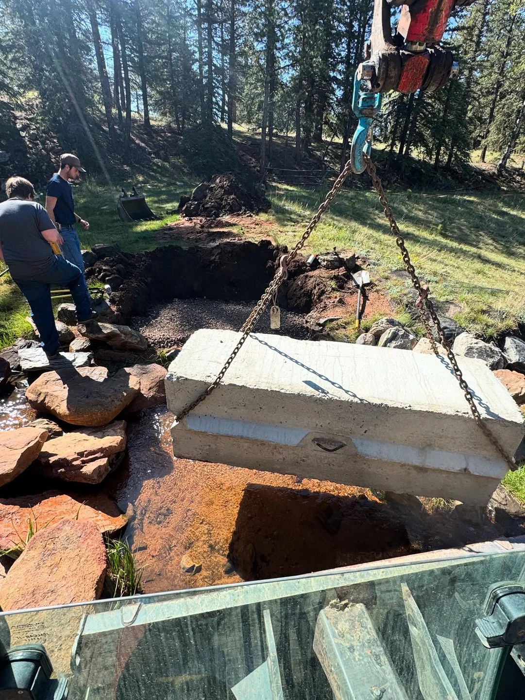 Crew guiding a concrete tank into an excavation among trees